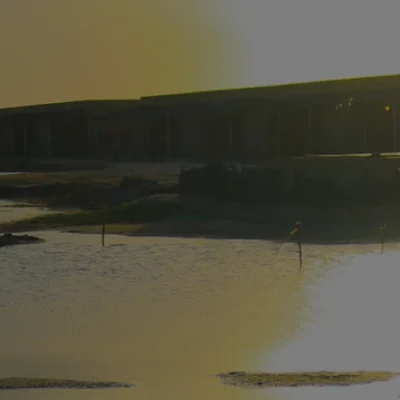 Vista al atardecer de una tranquila masa de agua con reflejos, siluetas de edificios y estructuras de madera a lo largo de la orilla, y un suave resplandor dorado en el cielo.