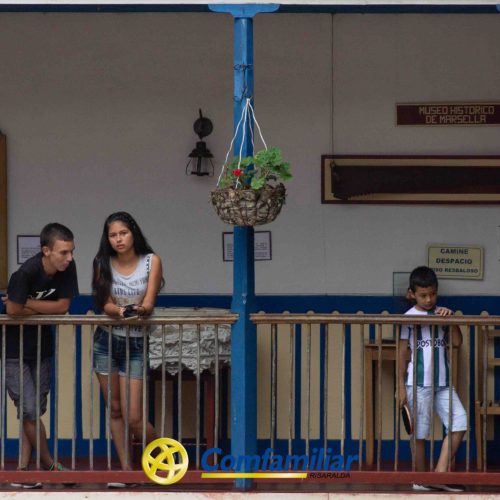 Tres personas están en un balcón azul y blanco de la Biblioteca Pública Municipal: un hombre y una mujer hablan a la izquierda, mientras que un niño está solo a la derecha. Plantas colgantes y carteles del museo León de Greiff decoran el fondo.