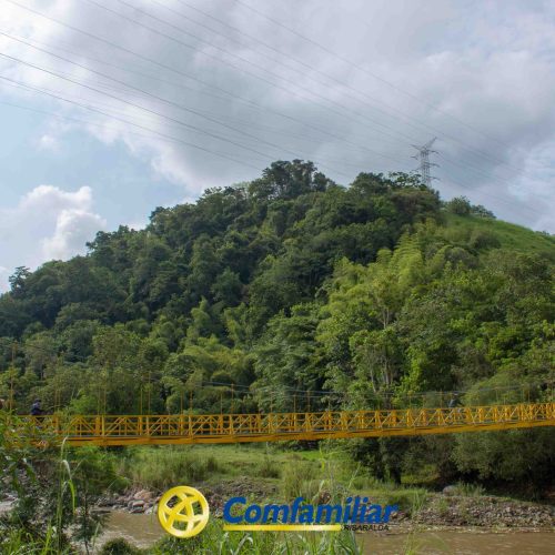 Un puente colgante amarillo cruza un río en un paisaje verde y exuberante cerca de Medellín, con una colina boscosa al fondo. El cielo está parcialmente nublado y el logotipo de Comfamiliar aparece en la parte inferior de la imagen.