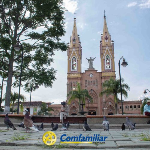 Una catedral con agujas gemelas se alza al fondo de una plaza de Medellín, donde la gente se sienta entre palomas y árboles. El logotipo y el texto de Comfamiliar aparecen al fondo, cerca de la famosa Biblioteca Pública León de Greiff.