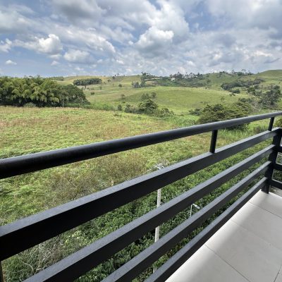 Disfrute de la serena vista desde el balcón del Mirador Campestre, que cuenta con una barandilla negra y baldosas de color claro, con vistas a exuberantes colinas verdes y árboles bajo un cielo nublado. Un pilar de madera a la izquierda añade encanto rústico a este tranquilo refugio.