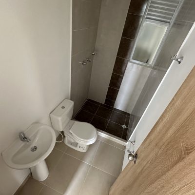 Pequeño cuarto de baño en el Mirador Campestre con lavabo blanco, inodoro y ducha acristalada. El suelo es de baldosas claras y la puerta, parcialmente abierta, presenta un acabado de madera. La ducha tiene azulejos más oscuros y una ventana esmerilada.