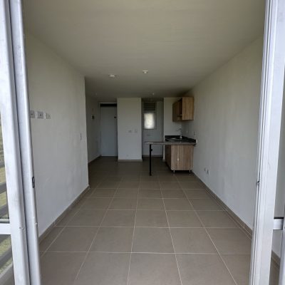 Vista de un pequeño apartamento sin amueblar del Mirador Campestre con suelos de baldosas, paredes blancas, una zona de cocina con armarios de madera y una gran puerta corredera de cristal que da al verde paisaje exterior.