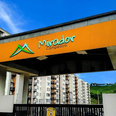 Puerta de entrada al complejo residencial Mirador Campestre, con señalización naranja, modernos edificios de apartamentos blancos y beige, y una ladera verde al fondo bajo un cielo azul.