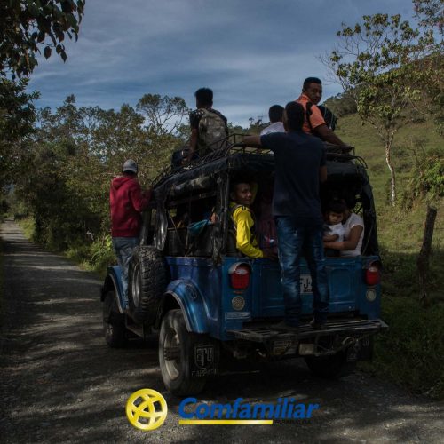 Un grupo de personas cabalga sobre y dentro de un jeep azul que circula por un camino rural de tierra en Mistrató, rodeado de vegetación y montañas, con el logotipo de Comfamiliar en la parte inferior de la imagen.