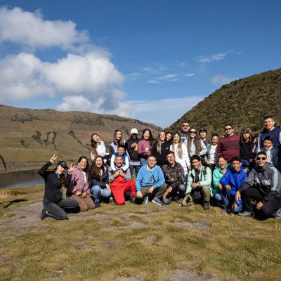Un grupo de unas 30 personas posan juntas al aire libre en una ladera cubierta de hierba cerca del Nevado del Ruiz, con montañas y un lago de fondo bajo un cielo azul. Todos sonríen y visten informalmente durante su pasadía por los termales.