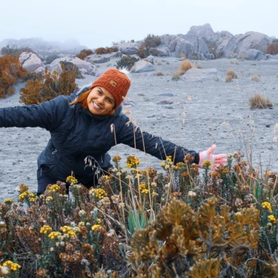 Una mujer con chaqueta negra, gorro naranja y guantes rosas sonríe con los brazos extendidos en medio del paisaje rocoso y brumoso del Nevado del Ruiz, con flores silvestres amarillas y arbustos en primer plano, perfecto para una aventura de pasadía.