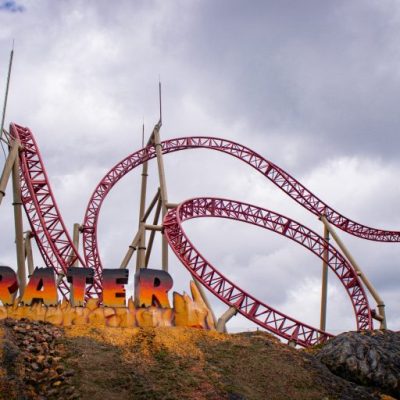 En el Parque del Café, una montaña rusa roja llamada Krater serpentea a través de bucles y giros bajo un cielo nublado, con su nombre escrito en negrita sobre una base rocosa pintada de amarillo en primer plano.