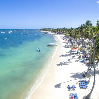 Vista aérea de una playa tropical de arena blanca, agua turquesa, palmeras, barcos en el océano y gente descansando en tumbonas bajo un cielo azul brillante.