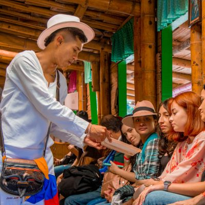 Un hombre vestido con ropa tradicional ofrece muestras de comida a un grupo de personas sentadas en un rústico escenario de madera. Algunas personas alcanzan la comida, mientras otras observan atentamente.