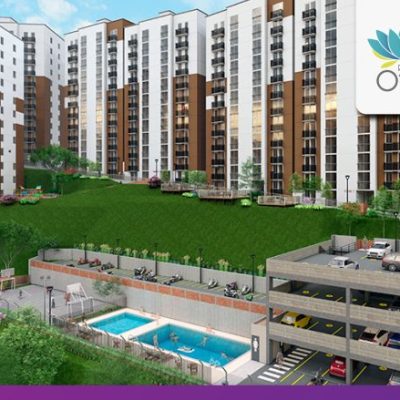 Un gran complejo moderno de apartamentos, Riveras del Otún, cuenta con varios edificios de gran altura blancos y marrones, césped verde, una zona de piscinas y un aparcamiento de varios niveles con varios coches aparcados. En la esquina aparece el logotipo de Otún.