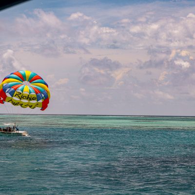 Un paravela de colores brillantes con la palabra Richie flota sobre una embarcación en aguas turquesas del océano bajo un cielo parcialmente nublado. Varias personas están en la barca y se ven arrecifes a lo lejos.