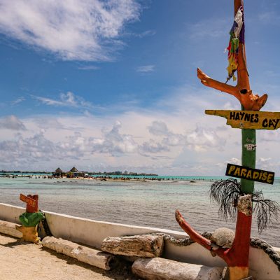 Un colorido cartel de madera en una playa de arena señala "Haynes Cay" y "Paradise", con aguas turquesas transparentes, gente nadando y una estructura con tejado de paja a lo lejos bajo un cielo azul con nubes.