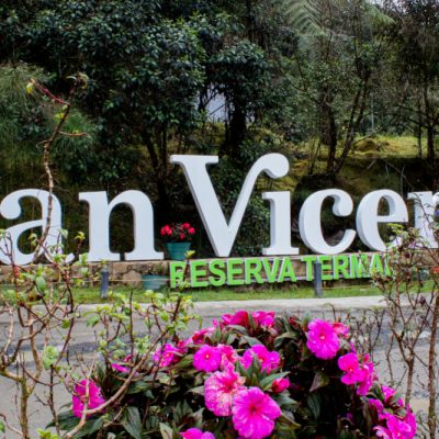 Unas grandes letras blancas que rezan "San Vicente" y unas letras verdes que rezan "Reserva Termal" le dan la bienvenida a Termales San Vicente, en medio de una exuberante vegetación y flores de colores.