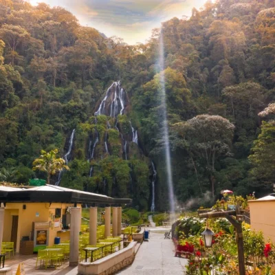 Una alta cascada cae por una exuberante ladera verde detrás de un patio con asientos al aire libre en Termales Santa Rosa, rodeado de árboles y plantas en flor bajo un cielo parcialmente nublado.