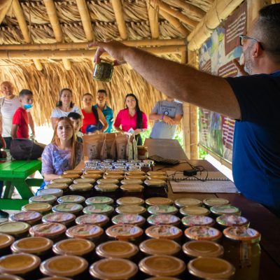 Un hombre en el Parque de la uva presenta tarros de productos a un grupo de personas atentas sentadas bajo un techo de paja, con tarros alineados sobre una mesa y carteles informativos en la pared detrás de él.